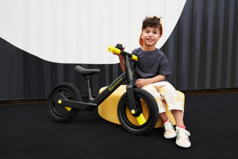 Un niño relajado está sentado en un puf en un espacio abstracto y sostiene una bicicleta de equilibrio negra con dos ruedas