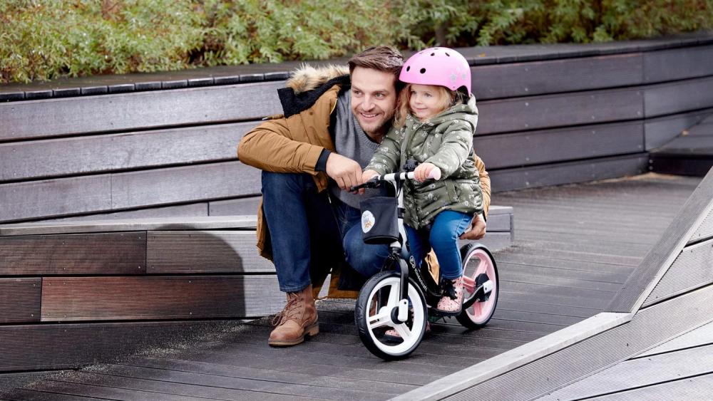Tiempo otoñal, un padre está afuera y se inclina sobre una niña que lleva puesto un casco y está sentada en una bicicleta de equilibrio con dos ruedas de colores negro y rosa