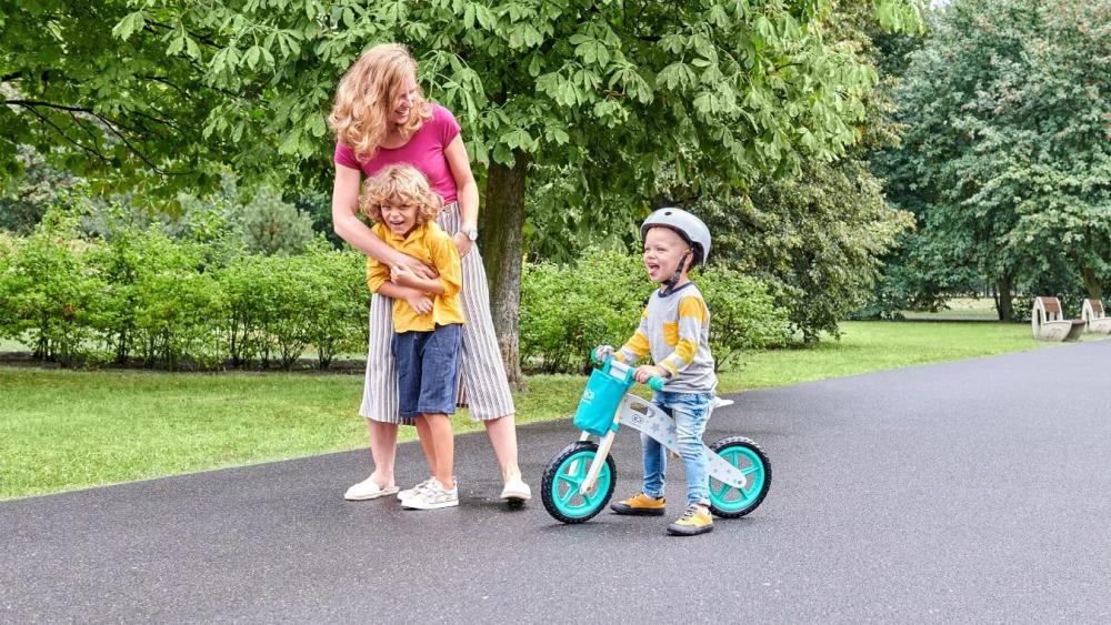 Una madre está paseando por el parque con su hijo en una bicicleta de equilibrio de dos ruedas de color turquesa y su otro hijo mayor