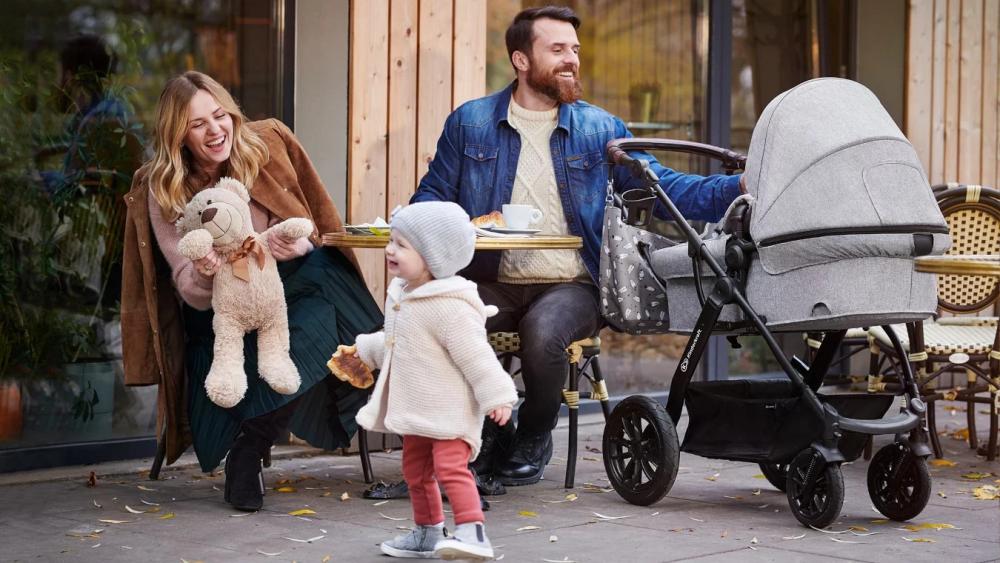 Mamá y papá en una cafetería al aire libre están jugando con un bebé que camina y un bebé acostado en un carrito profundo de color gris