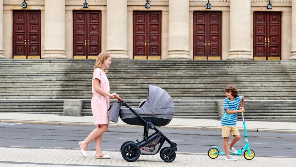 Una madre está paseando por la ciudad con un carrito profundo de color gris y un bebé en un patinete de dos ruedas de color turquesa