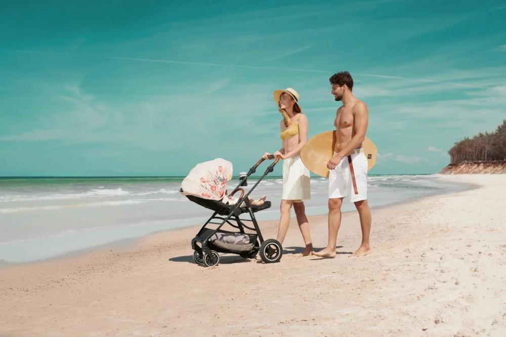 Mamá y papá están paseandose por la playa con un bebé tumbado en una silla de paseo negra con cabina beige