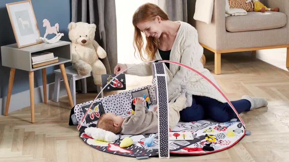Una madre sonriente está en u n apartamento y juega con un bebé tumbado en un tapete sensorial de colores mostrándole una imagen de contraste
