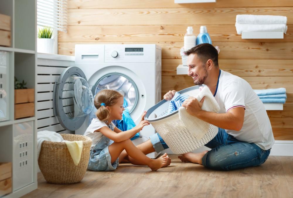 Un padre sonriente le muestra a su hija una cesta de la colada, lo están pasando bien junto a la lavadora