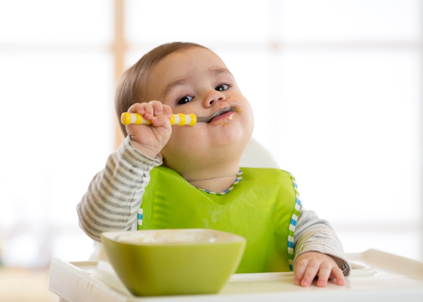 Un niño está sentado en una trona para bebés, delante tiene un cuenco verde y lleva puesto un babero verde. Está comiendo con una cuchara, mirando a la cámara