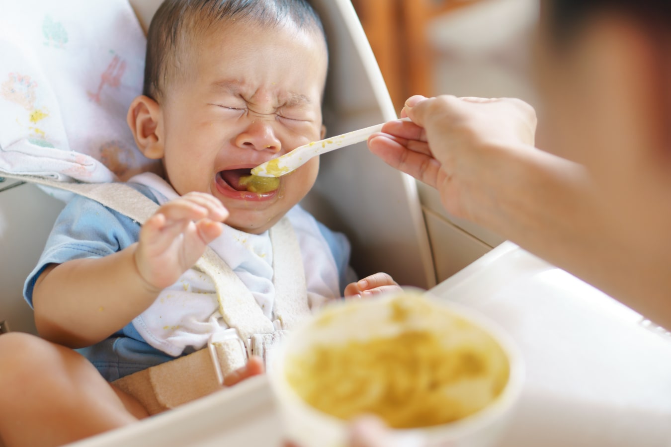 Un niño está sentado en una trona para bebés, se niega a comer con una cuchara y está llorando.