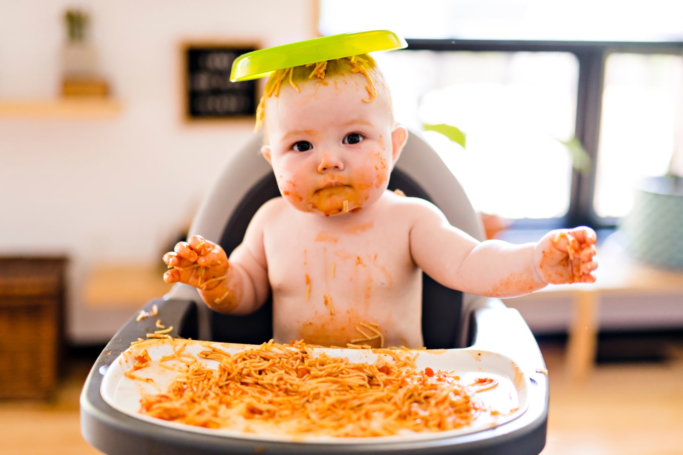 Un niño ensuciado con un espagueti está sentado en una trona para bebés, en la cabeza tiene un plato con la comida