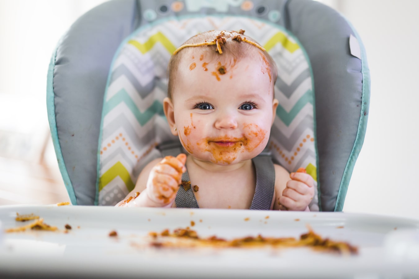 Un niño ensuciado con un espagueti está sentado en una trona para bebés. Está feliz y sonriente, hay mucha comida sobre él y sobre la bandeja.