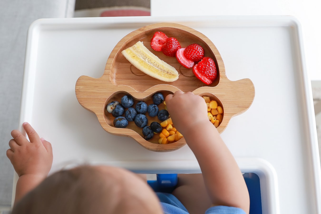 En un plato de madera con compartimentos hay frutas y verduras de colores: plátano, arándanos, maíz y fresas. El niño estira la mano para coger la comida