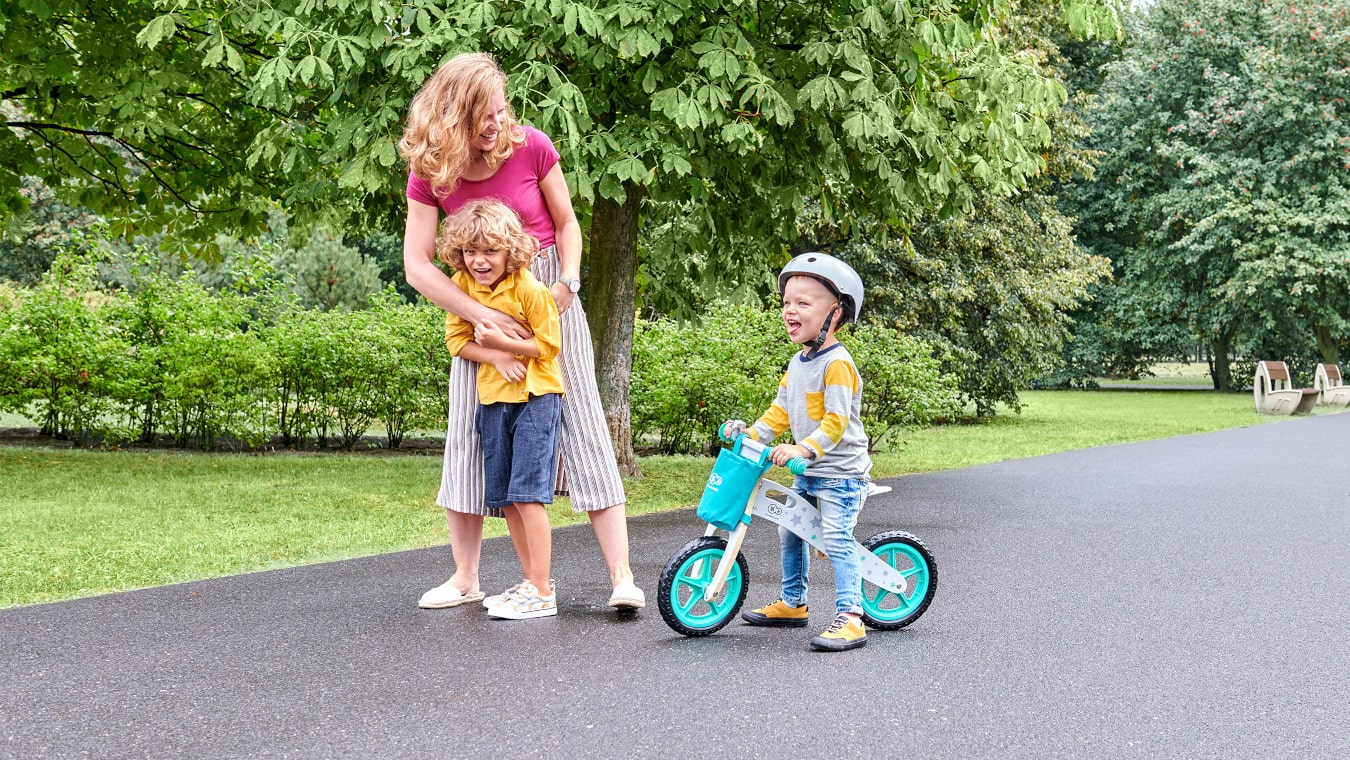 Tiempo otoñal, un padre está afuera y se inclina sobre una niña que lleva puesto un casco y está sentada en una bicicleta de equilibrio con dos ruedas de colores negro y rosa