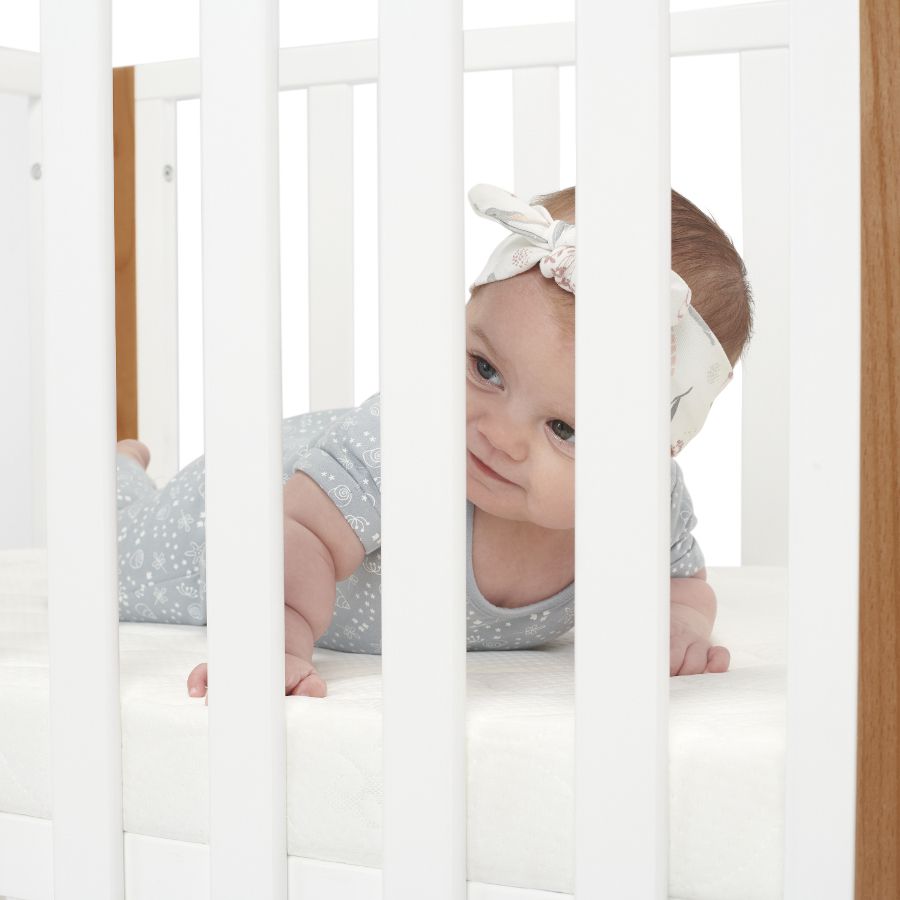Un bebé de varios meses está acostado en un cómodo colchón y mira a través de los barrotes blancos de una cuna de la marca Kinderkraft.