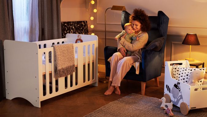 Por la noche, en una habitación de bebé, una madre está sentada en un sillón y sostiene a un bebé. A su lado, hay un parque de juegos y un baúl para los juguetes Racoon de la marca Kinderkraft.