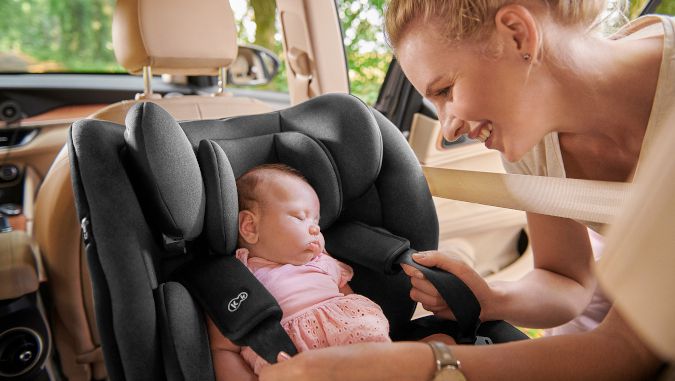 Dentro de un coche, una madre y dos niños están sentados en unas sillas de coche de Kinderkraft. El recién nacido duerme orientado hacia atrás y la madre se voltea para ver al niño mayor.