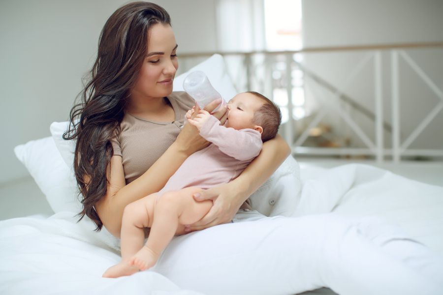En una casa, una madre está sentada en su cama y sujeta a un bebé en brazos. Lo mira sonriendo y lo alimenta con un biberón.