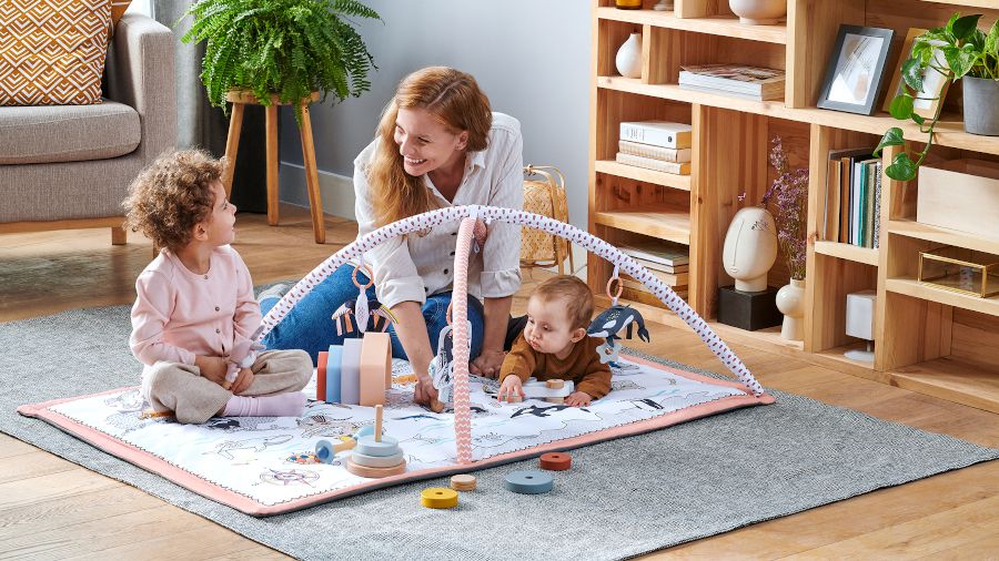 En un apartamento, sobre la alfombra está extendido un tapete de la marca Kinderkraft. Una madre, un niño y un bebé están jugando encima del tapete.
