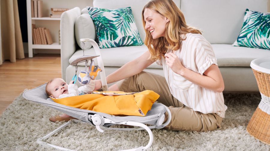 En una habitación hay un bebé acostado en una silla mecedora con un carrusel con juguetes colgado encima. Al lado está sentada su mamá que está mirando al niño.