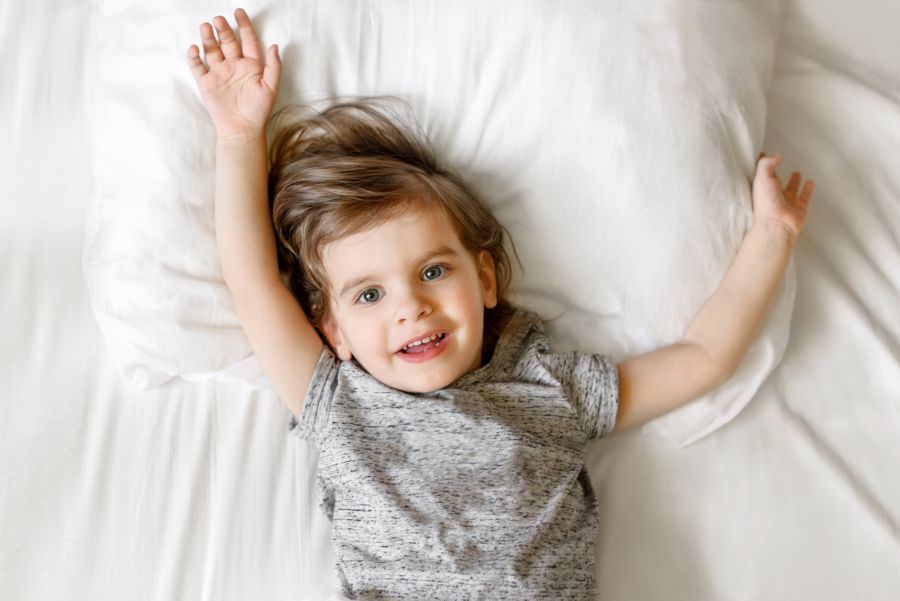 Un niño está acostado boca arriba en una cama con sábanas blancas. Mira al objetivo y extiende los brazos hacia los lados mostrando que está cómodo.