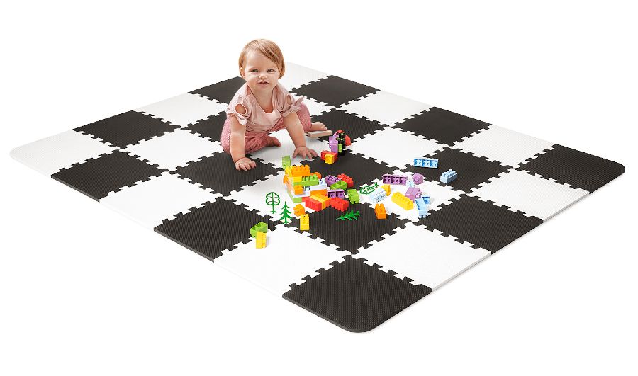Sobre el fondo blanco hay un rompecabezas blando blanco y negro de la marca Kinderkraft. Encima de él está sentada una niña que juega con cubos.