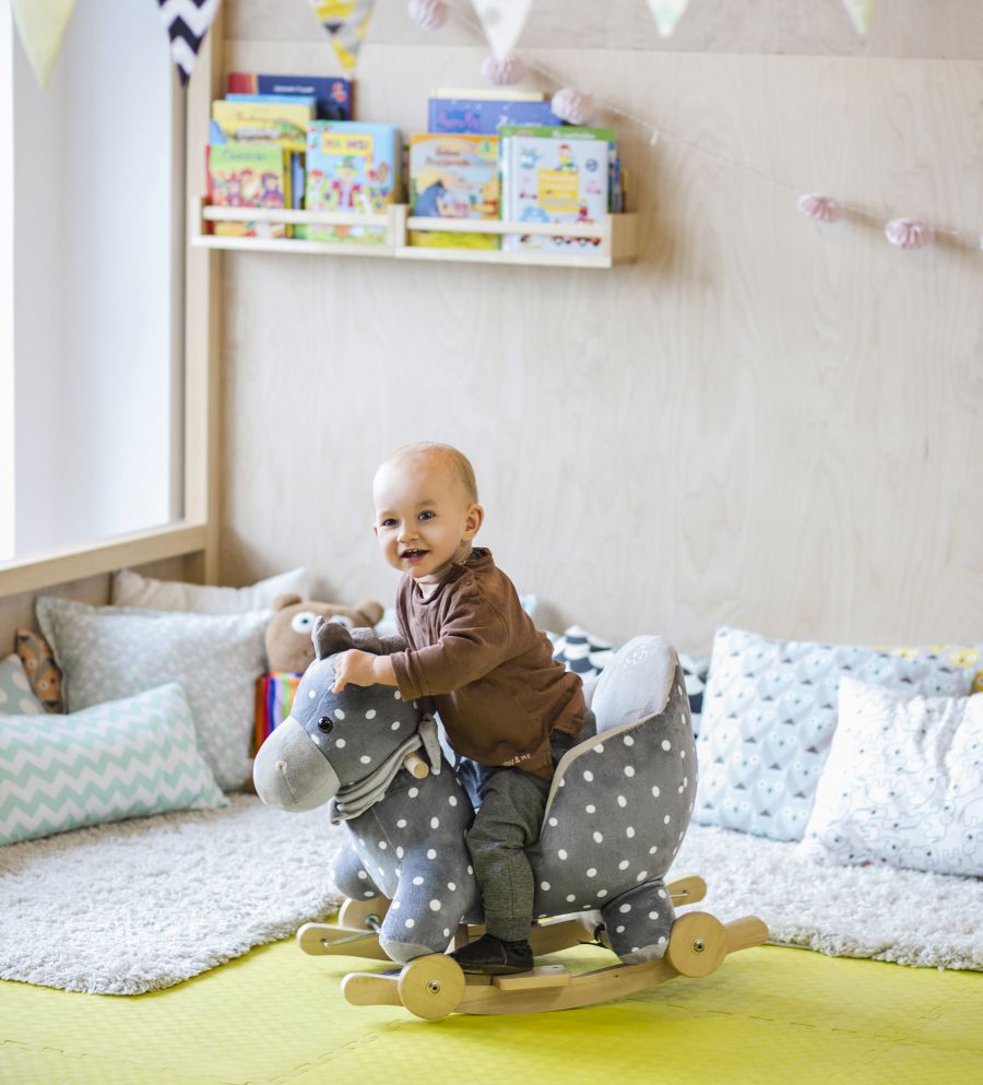 En una habitación infantil, un bebé sonriente está sentado en un caballito balancín.