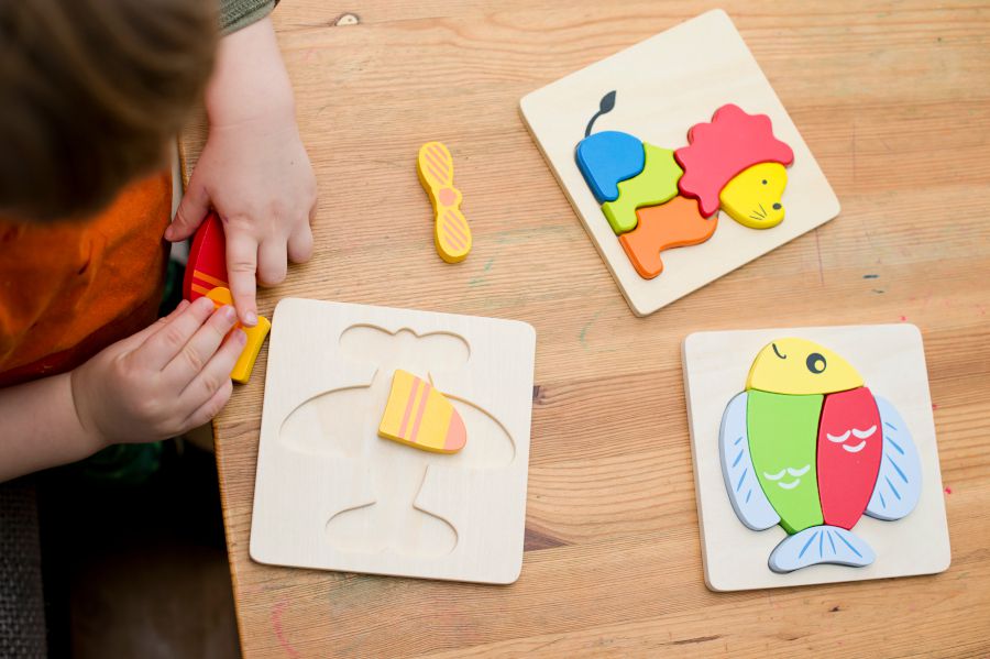 Un niño está colocando sobre la mesa unas piezas de un rompecabezas de madera con dibujos de un león, un pez y un avión.