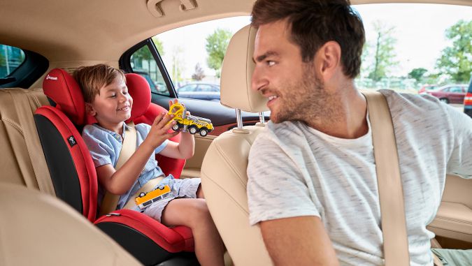 Dentro de un coche, un padre-conductor se voltea atrás para ver a su niño pequeño que está jugando sentado en una silla de coche de la marca Kinderkraft.