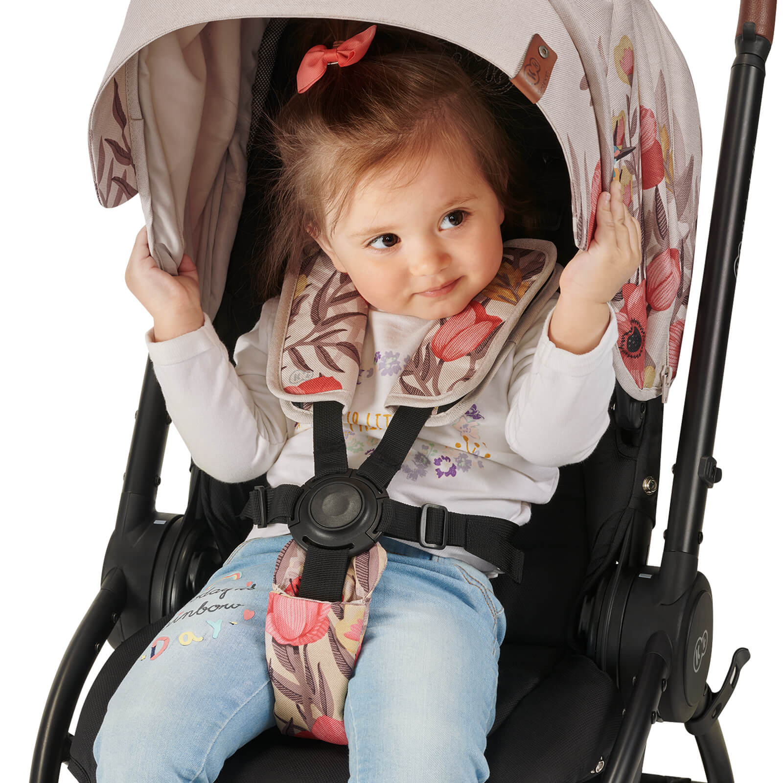 Una niña pequeña está sentada en la silla de paseo, abrochada con cinturones, agarra la visera y sonríe