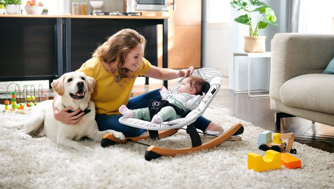 En un apartamento, un bebé está acostado en una silla mecedora de la marca Kinderkraft. Al lado está su mamá que acaricia la cabeza del niño con una mano y abraza a un perro grande con la otra.