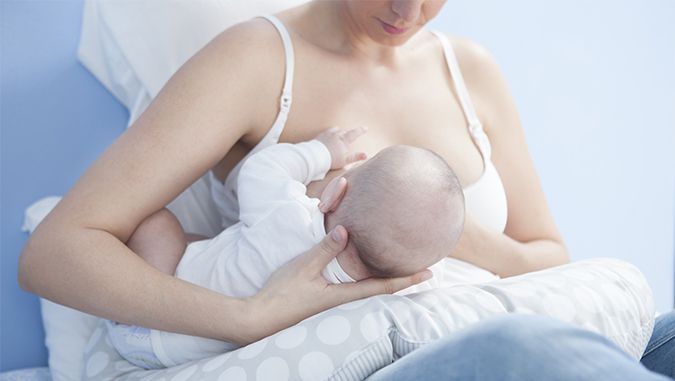 Madre está sentada en una cama con la espalda apoyada contra la pared y da de comer a su bebé acostado en una almohada de lactancia especial de la marca Kinderkraft.