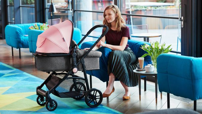 Madre vestida elegante está sentada en una cafetería y mira hacia un carrito de bebé gris: una góndola de la marca Kinderkraft, en la que está acostado su bebé.