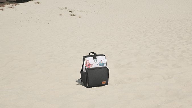 Es un día soleado, en una playa de arena hay una mochila blanca y gris de la marca Kinderkraft con motivos de flores y pájaros.