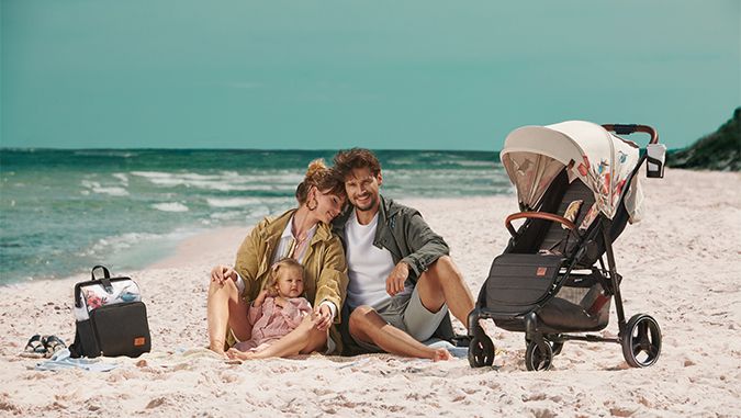 Una niña y sus padres están sentados en una playa. Junto a ellos hay una mochila de la marca Kinderkraft con motivos de flores y pájaros y un carrito para bebé con el mismo diseño.