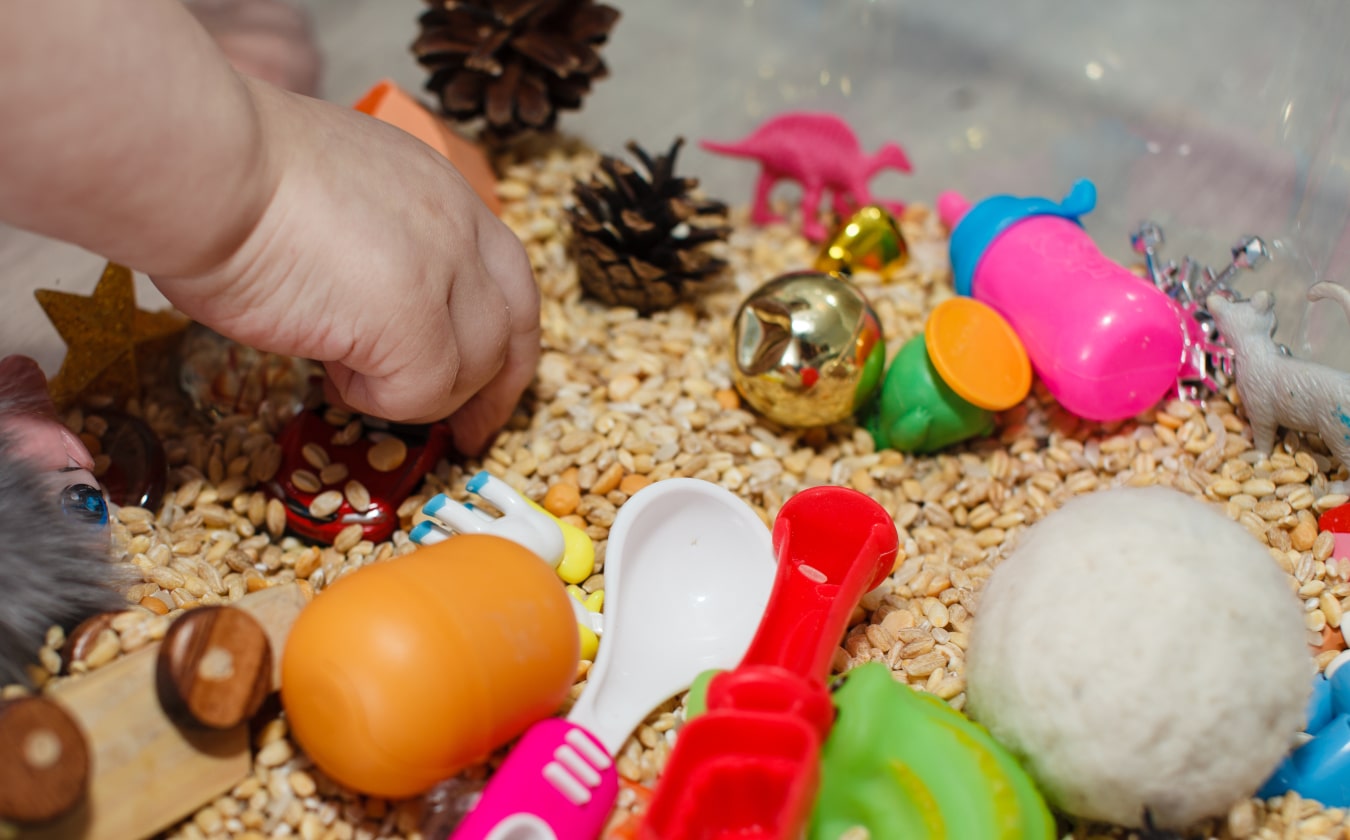 la mano del niño toca granitos en los que hay juguetes infantiles de colores. El peque conoce objetos de nuevas texturas mediante el tacto