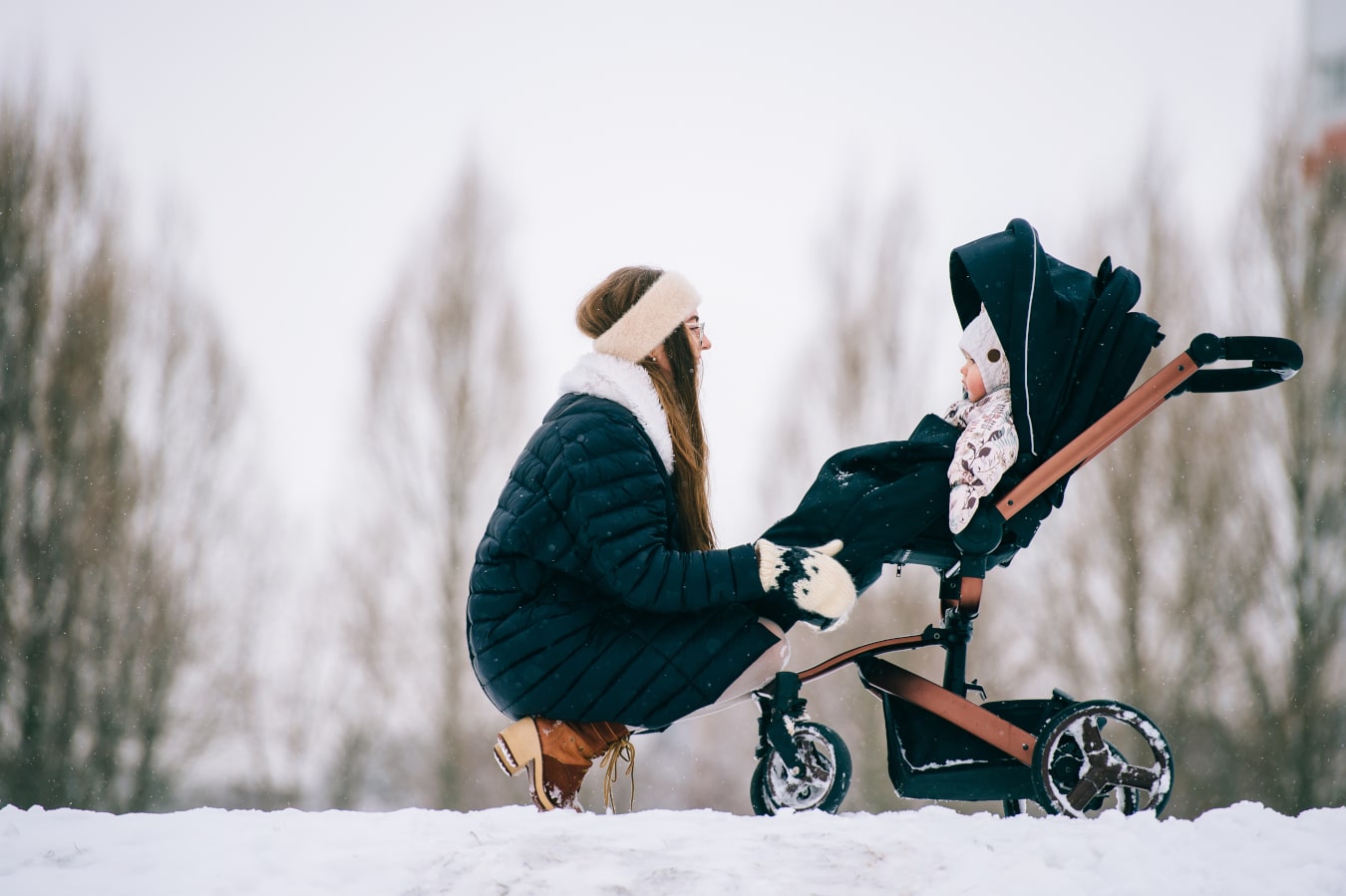 Es invierno, la madre se agacha junto a la silla de paseo y se inclina para ver al niño. Hay mucha nieve, todos llevan chaquetas