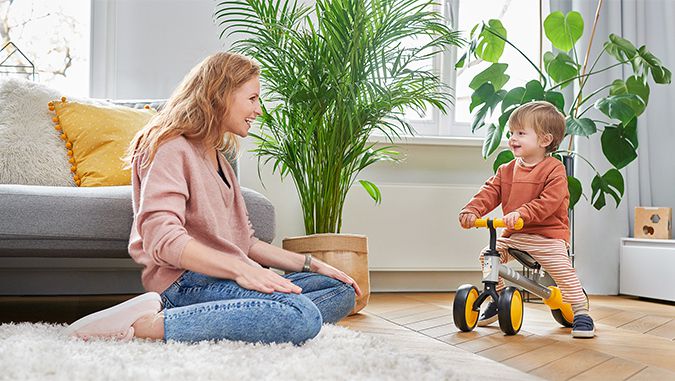 Un niño se está paseando por una habitación en un triciclo de equilibrio de la marca Kinderkraft. Su mamá está sentada al lado en el suelo y lo está observando con una sonrisa.