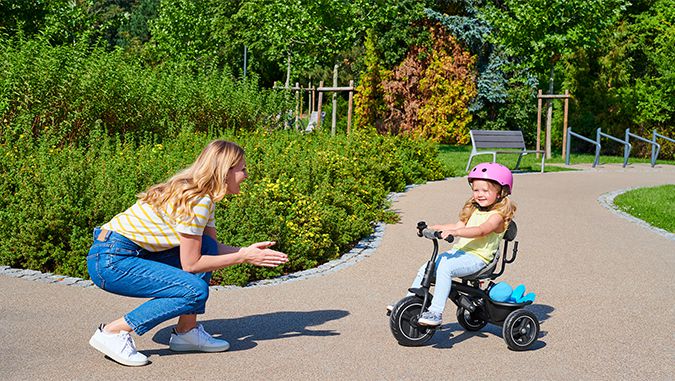 Es un día soleado, en un sendero de un parque una niña con un casco rosa va montando su triciclo de la marca Kinderkraft en la dirección de su madre sonriente.