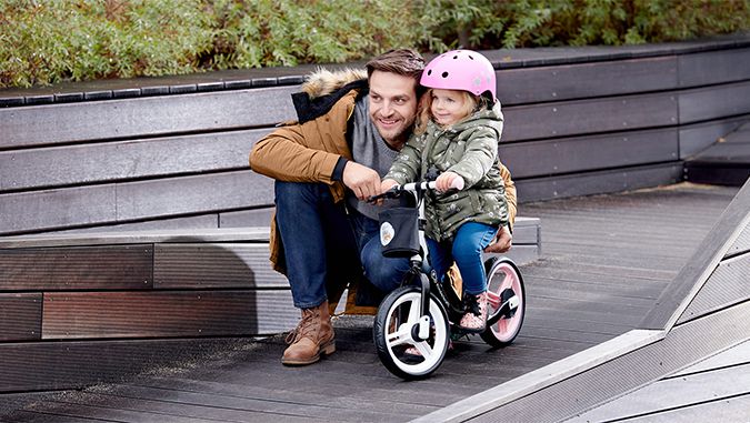 Una niña con un casco rosa está sentada en una bicicleta de la marca Kinderkraft. Su padre la apoya sujetando es asiento. Ambos miran en la misma dirección.