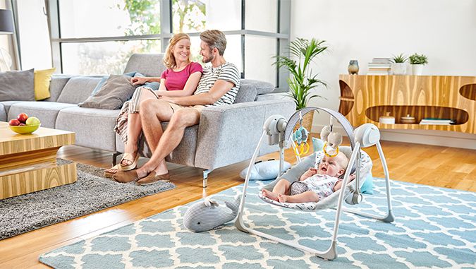 En un sofá, unos padres sonrientes se están mirando mutuamente. Junto a ellos, en la alfombra hay una silla mecedora de la marca Kinderkraft en la que está acostado un bebé viendo los juguetes colgados encima.