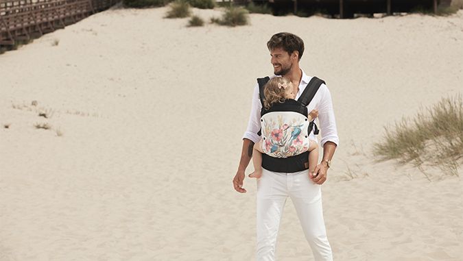 Padre vestido con una camisa y pantalón blanco elegante está en una playa. En su barriga lleva un portabebés de la marca Kinderkraft en el que está sentada una niña.
