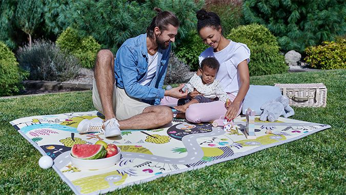 Es un día soleado, unos padres sonrientes están sentados en la hierba y juegan con un bebé. Junto a ellos hay juguetes, un plato de fruta y una cesta de picnic.