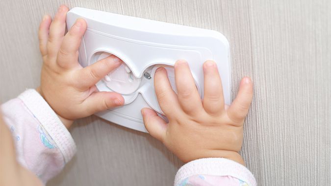 Las manos de un niño están apoyadas contra el enchufe. El niño intenta meter los dedos dentro, pero el enchufe tiene dispositivos de seguridad que lo impiden.