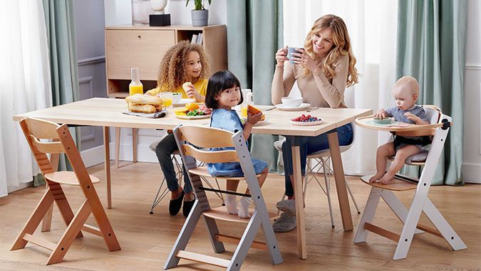 Madre y tres hijos desayunan sentados a la mesa. Dos niños más pequeños están sentados en unas sillas especiales de la marca Kinderkraft.