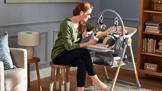 Madre sonriente está sentada en una pequeña silla en una habitación. Sostiene el pie de un bebé que está durmiendo a su lado en una silla de la marca Kinderkraft.