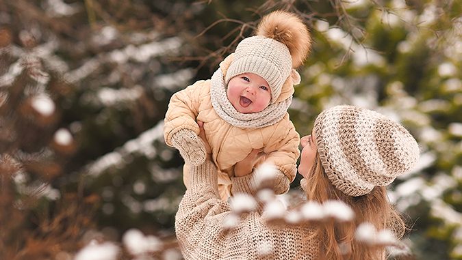 Invierno, se puede ver la nieve en los árboles del fondo. Madre viste un jersey de lana y un gorro, levanta a un niño sonriente con un gorro y mono de invierno.