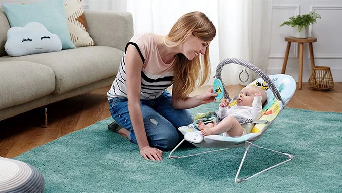 Madre se arrodilla en la alfombra y, con una sonrisa, muestra un juguete a su bebé que está acostado en la silla mecedora-hamaca de la marca Kinderkraft. El niño lo observa con interés..