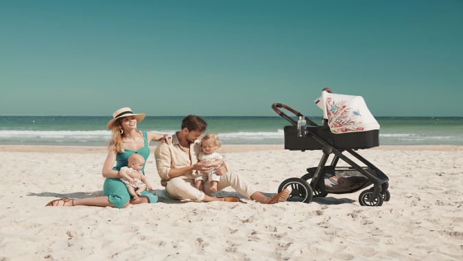 Es un día soleado, unos padres están sentados en una playa con dos niños pequeños. Al lado hay un carrito de bebé: una góndola de la marca Kinderkraft con una cabina de colores. 