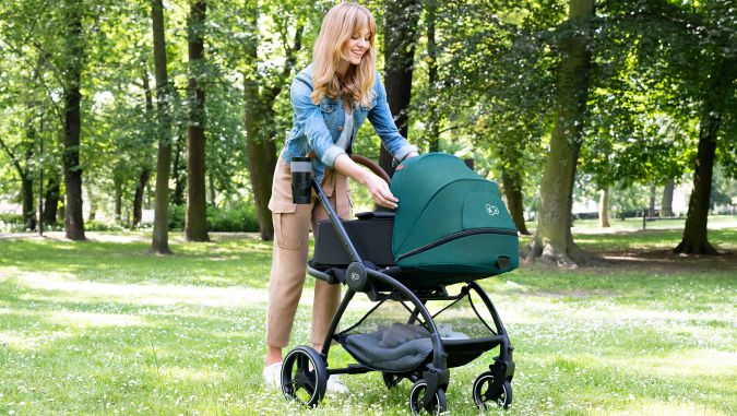 Madre sonriente está en un parque sobre la hierba y despliega una cabina del carrito de bebé verde: una góndola de la marca Kinderkraft.