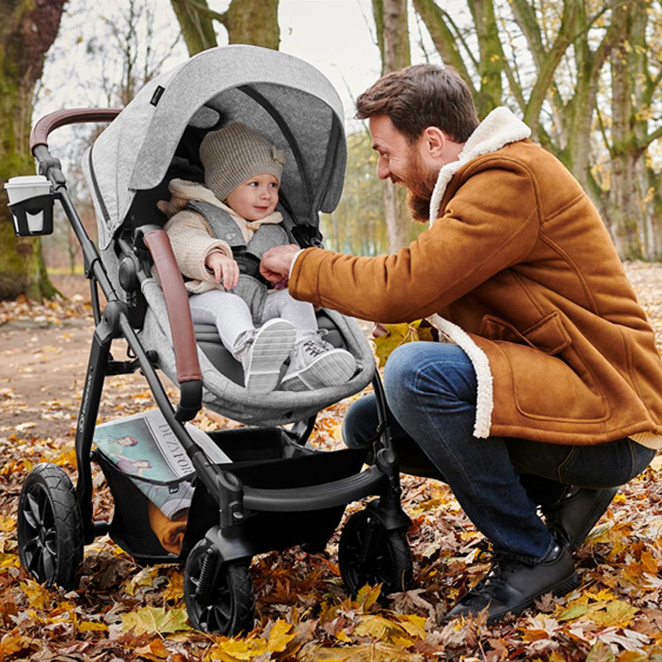 El  padre vestido de chaqueta se inclina hacia un niño sentado en un carrito de bebé. El niño sonríe, hay hojas de otoño alrededor