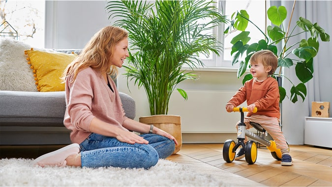 La madre sonríe a su hijo montado en el triciclo cutie kinderkraft. Están en casa, al fondo se ven plantas verdes y un sofá gris. El niño sonríe.