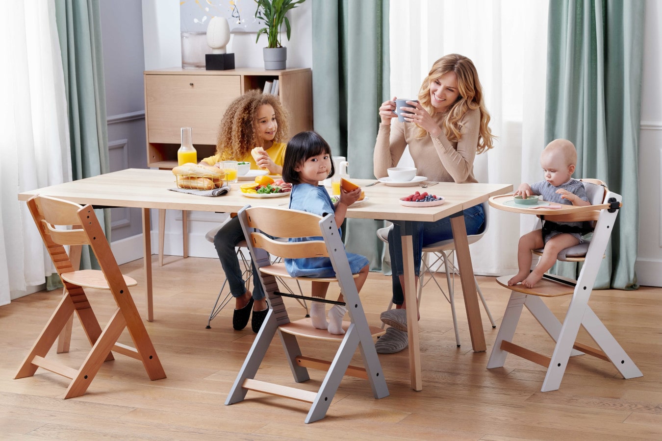 Mamá y tres niños están sentados en una mesa, la mamá está tomando un café, los niños están contentos. La trona para bebés ENOCK dispone de 3 funciones: desde una trona para bebés, hasta un sillón para niños mayores.
