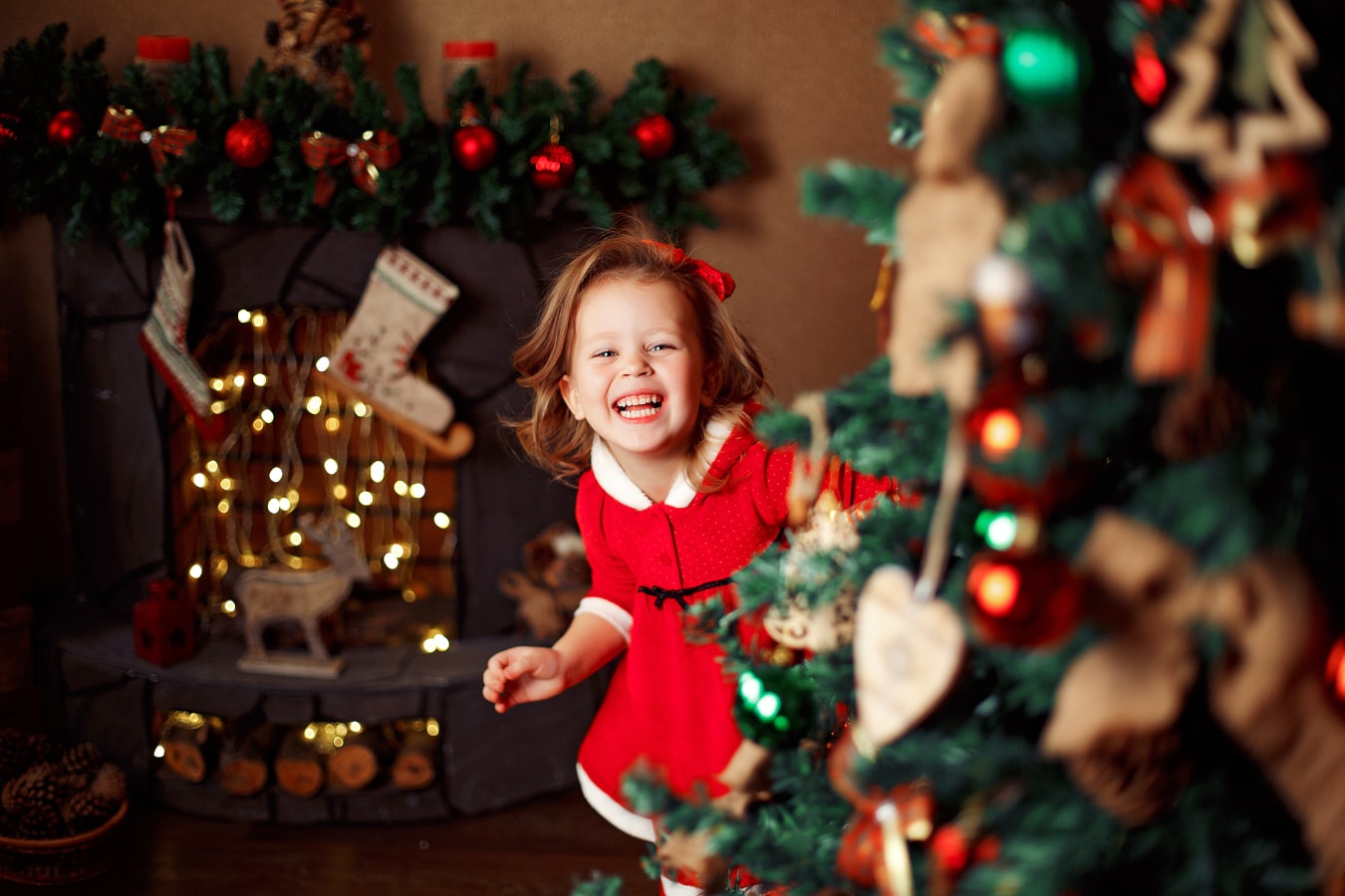 Una niña alegre con un vestido rojo se asoma por detrás del árbol de Navidad, está riéndose, está feliz.